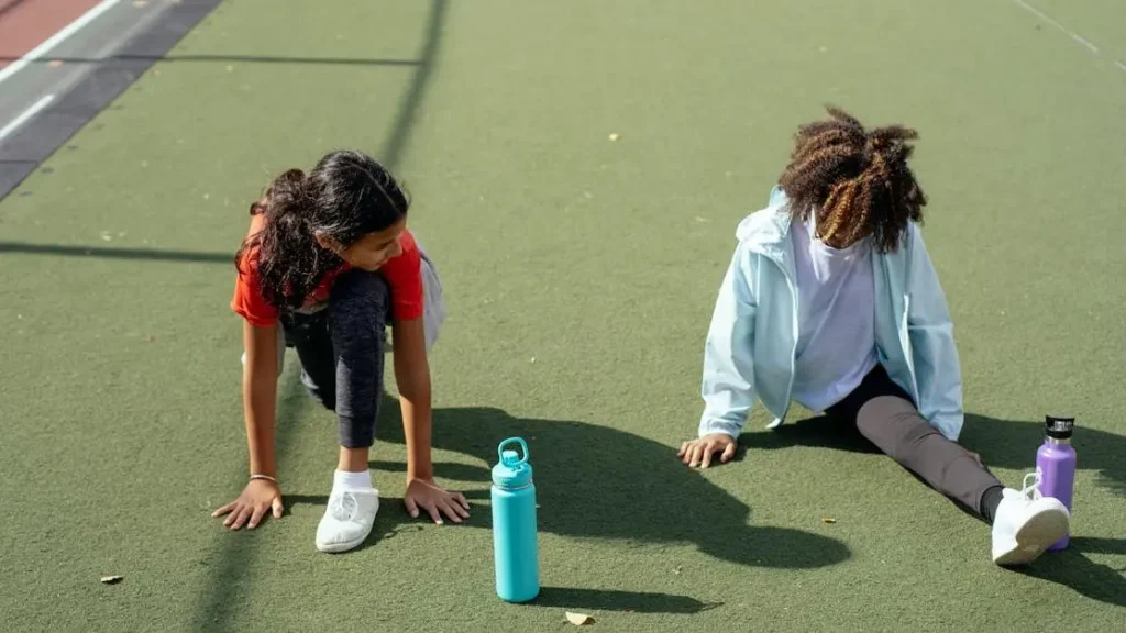 Black girl performing split while friend stretches before calisthenics exercises at stadium